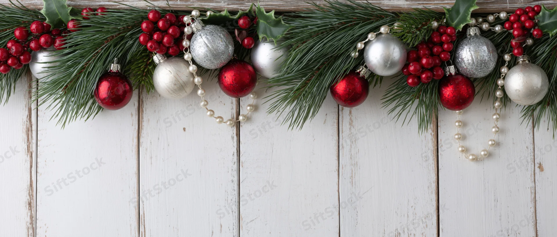 Christmas garland with pine branches, holly, red and silver ornaments, and pearl beads arranged on a white wooden background