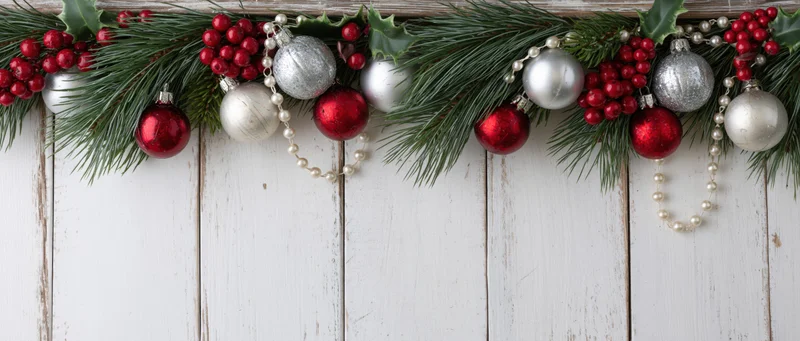 Christmas garland with pine branches, holly, red and silver ornaments, and pearl beads arranged on a white wooden background