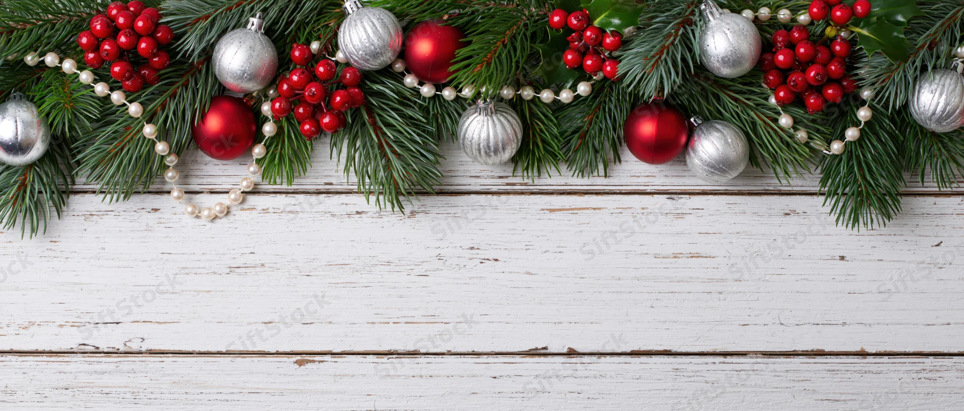 Christmas garland with pine branches, red and silver ornaments, berries, and pearls arranged along white weathered wood surface with copyspace below
