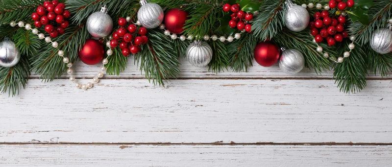Christmas garland with pine branches, red and silver ornaments, berries, and pearls arranged along white weathered wood surface with copyspace below