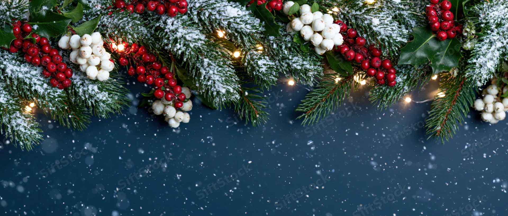 Snowy evergreen garland with red and white berries and warm lights arranged across dark blue background with falling snow and copyspace below