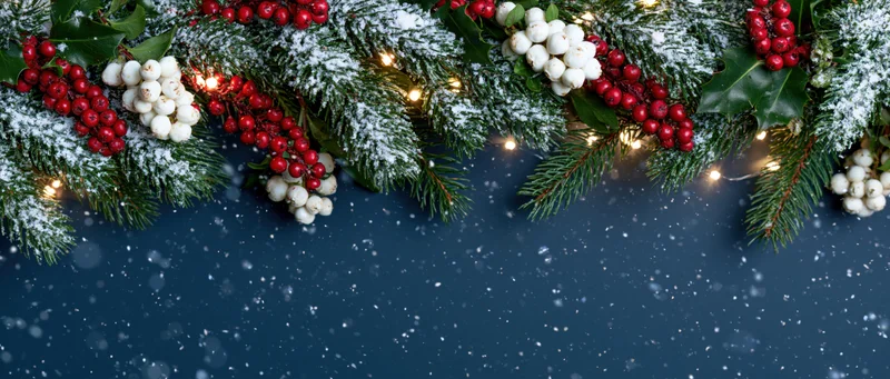 Snowy evergreen garland with red and white berries and warm lights arranged across dark blue background with falling snow and copyspace below
