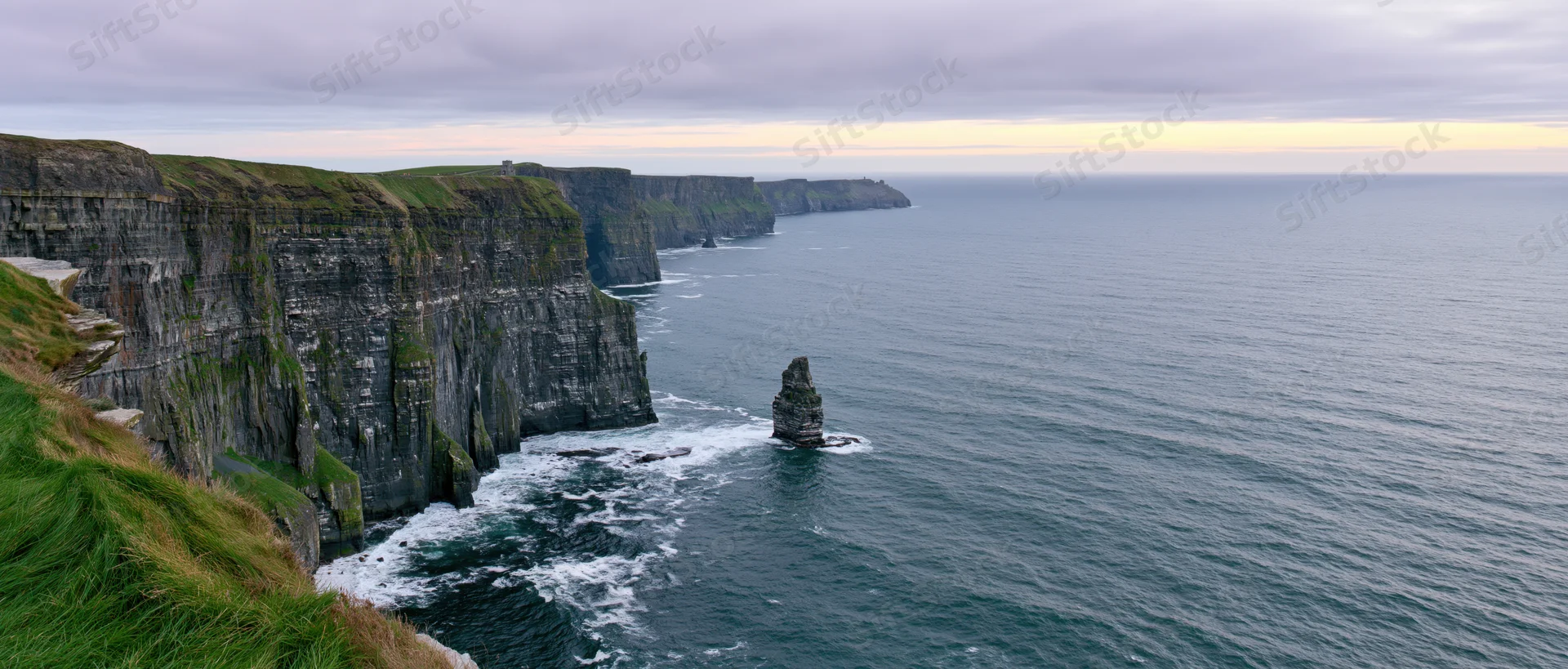 Rugged cliff face overlooking the ocean with sea stacks and grassy top under overcast sky