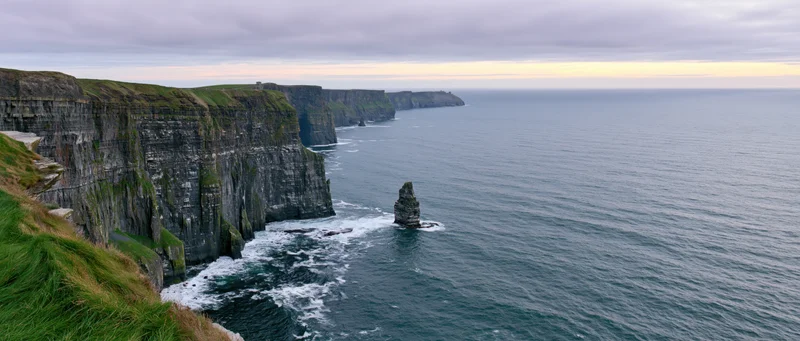 Rugged cliff face overlooking the ocean with sea stacks and grassy top under overcast sky
