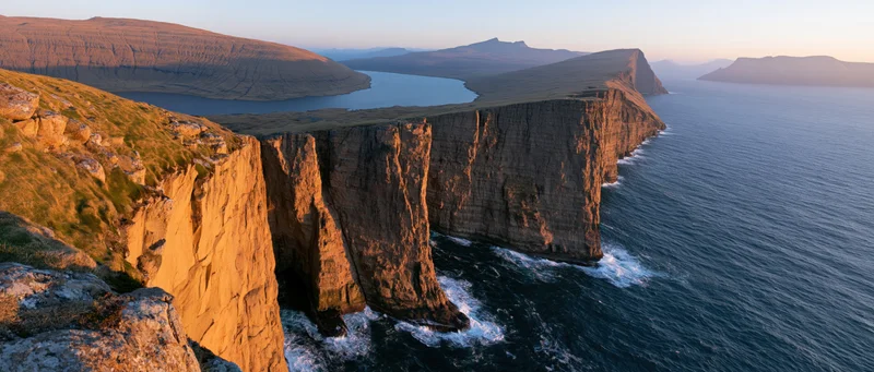 Rugged cliff face overlooking the ocean at sunset with layered rock formations and crashing waves along the coastline