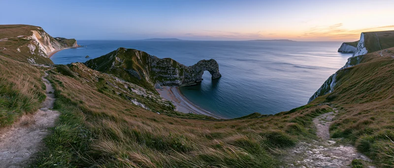 Natural rock arch on a coastal cliff overlooking a calm sea at sunset, with a winding path through grassy hills