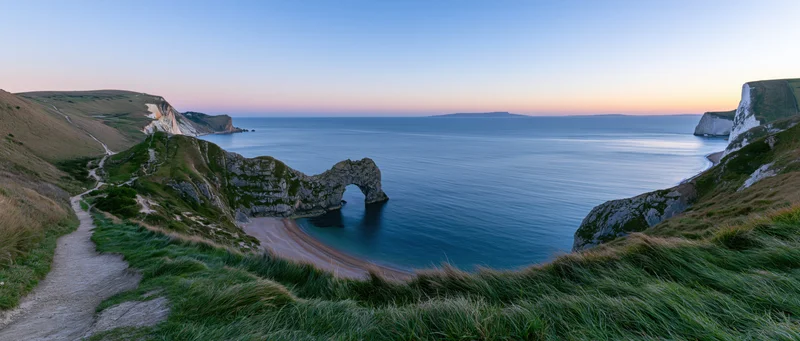 Natural rock arch on a coastal cliff overlooking a calm sea at sunset with a winding path through grassy terrain