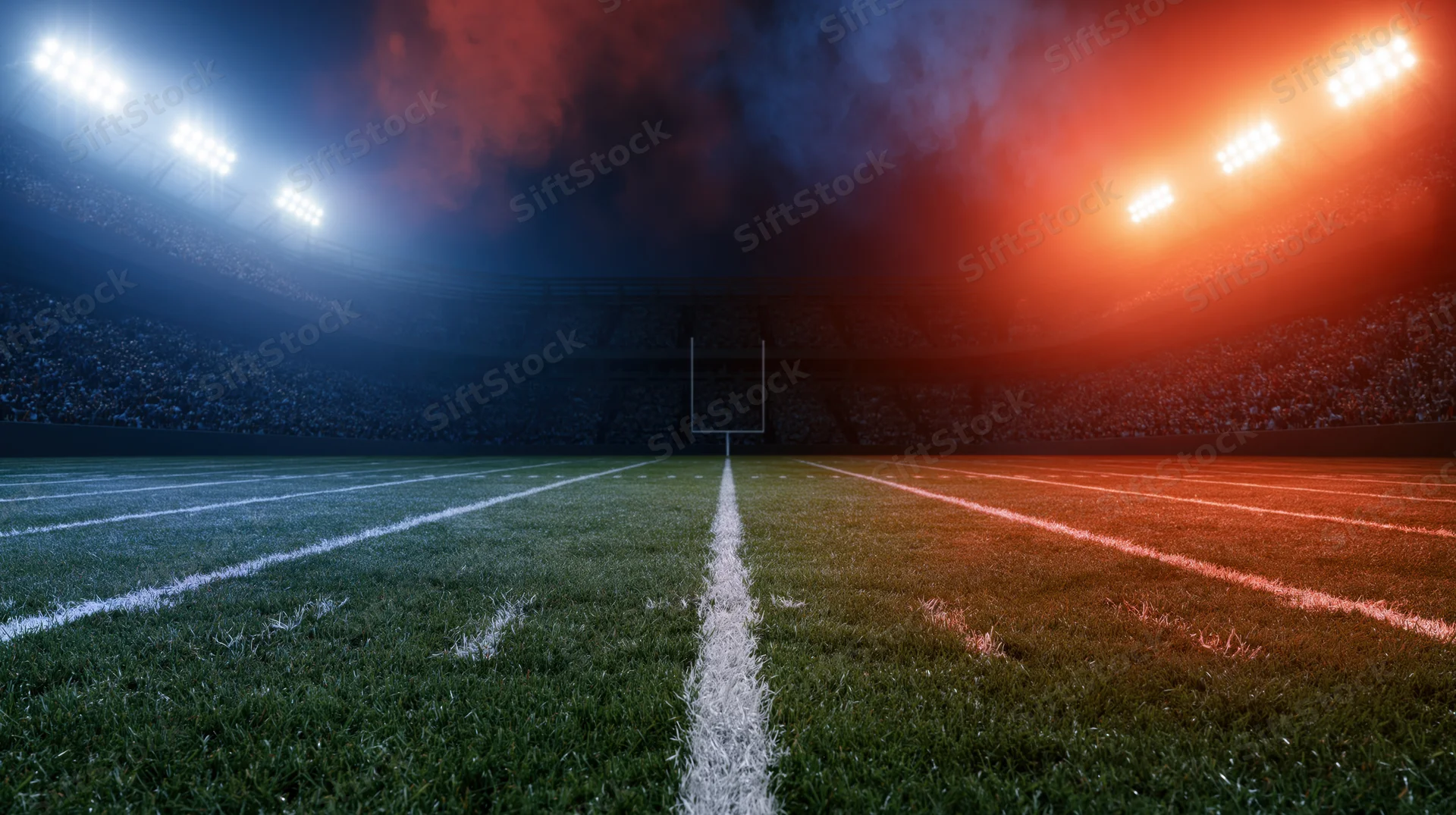 Football field at night under red and blue stadium lights with goalpost at center and crowd in stands