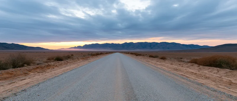 Gravel road stretching into desert landscape toward distant mountains at sunset