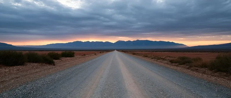 Straight gravel road stretching through arid desert toward distant mountains at sunset