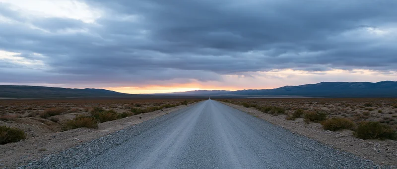 Gravel road stretching into the desert toward distant mountains under a cloudy sunset sky