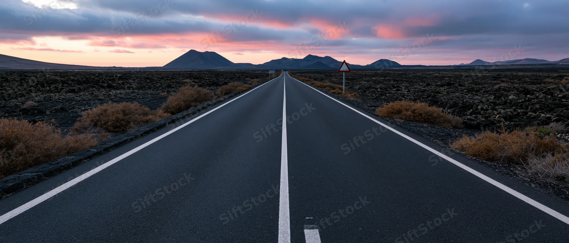 Straight asphalt road stretching toward distant mountains under a pink and orange sunset sky