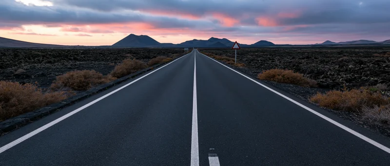 Straight asphalt road stretching toward distant mountains under a pink and orange sunset sky