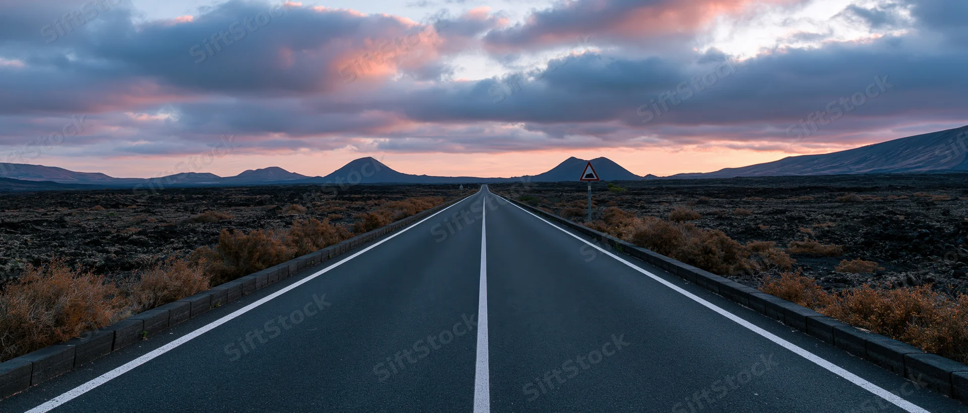 Straight asphalt road stretching toward distant volcanic mountains under a colorful twilight sky in a barren lava desert landscape