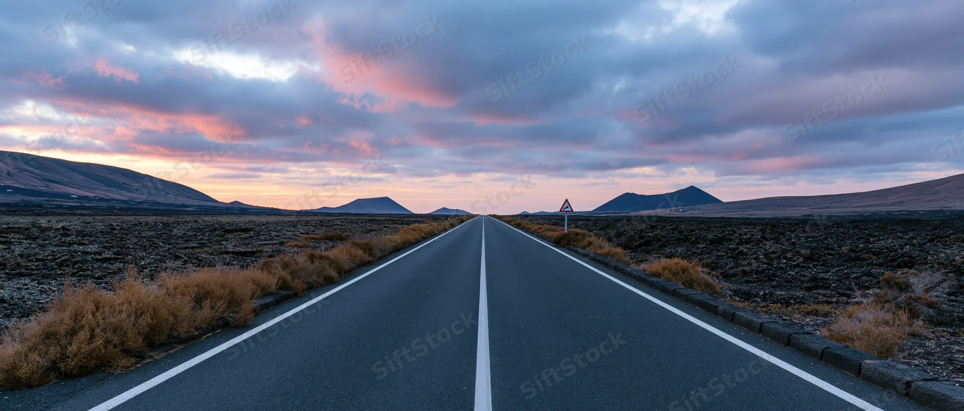 Straight asphalt road stretching toward distant volcanic mountains under a pink and orange twilight sky