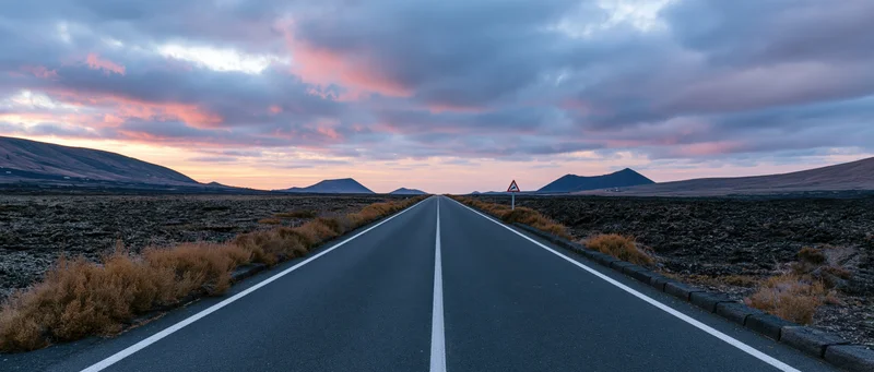 Straight asphalt road stretching toward distant volcanic mountains under a pink and orange twilight sky