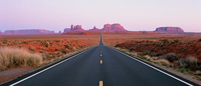 Straight asphalt road stretching into the desert toward red rock mesas at twilight