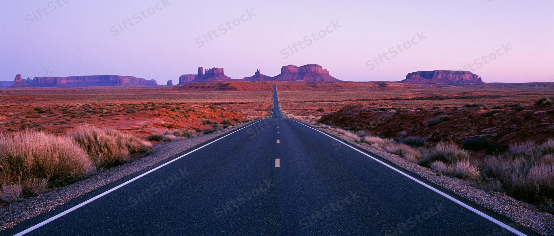 Straight asphalt highway stretching into the desert landscape with red rock mesas under twilight sky