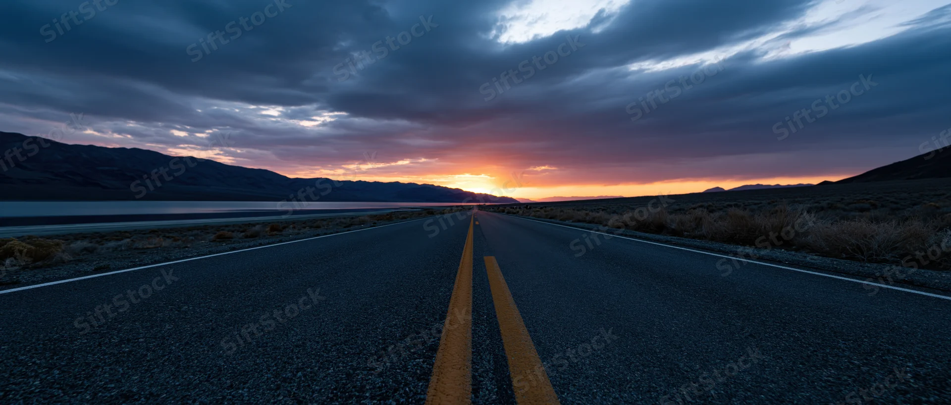 Straight asphalt road stretching toward a sunset horizon in a desert landscape with mountains and shrubs on either side