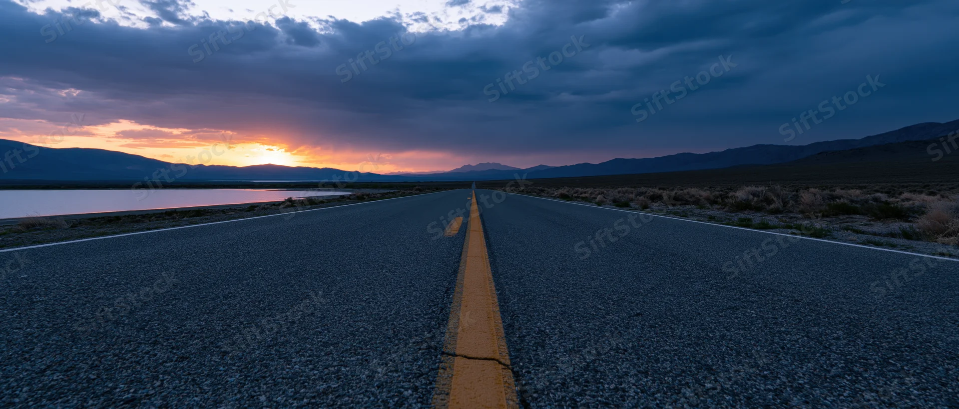 Straight asphalt road stretching toward distant mountains at sunset beside a calm lake