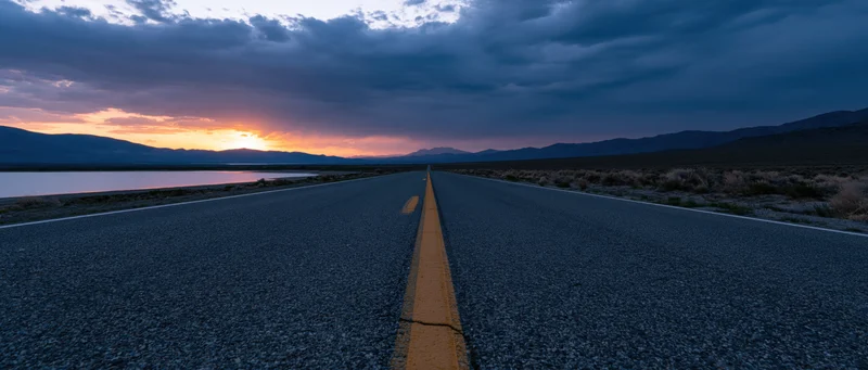 Straight asphalt road stretching toward distant mountains at sunset beside a calm lake