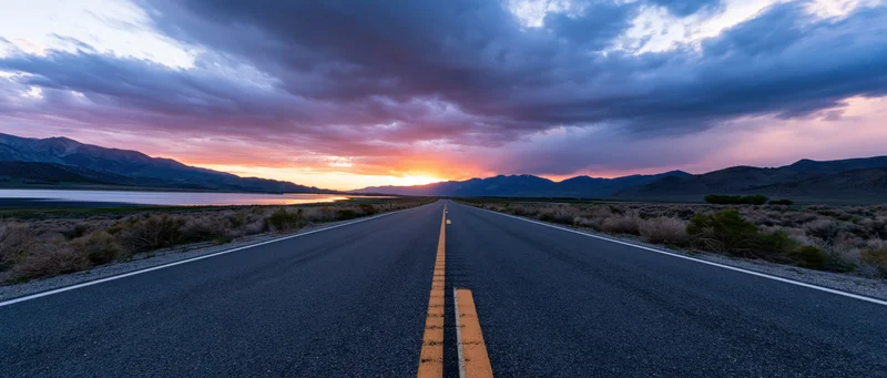 Straight asphalt road leading toward sunset over mountains and lake in desert landscape