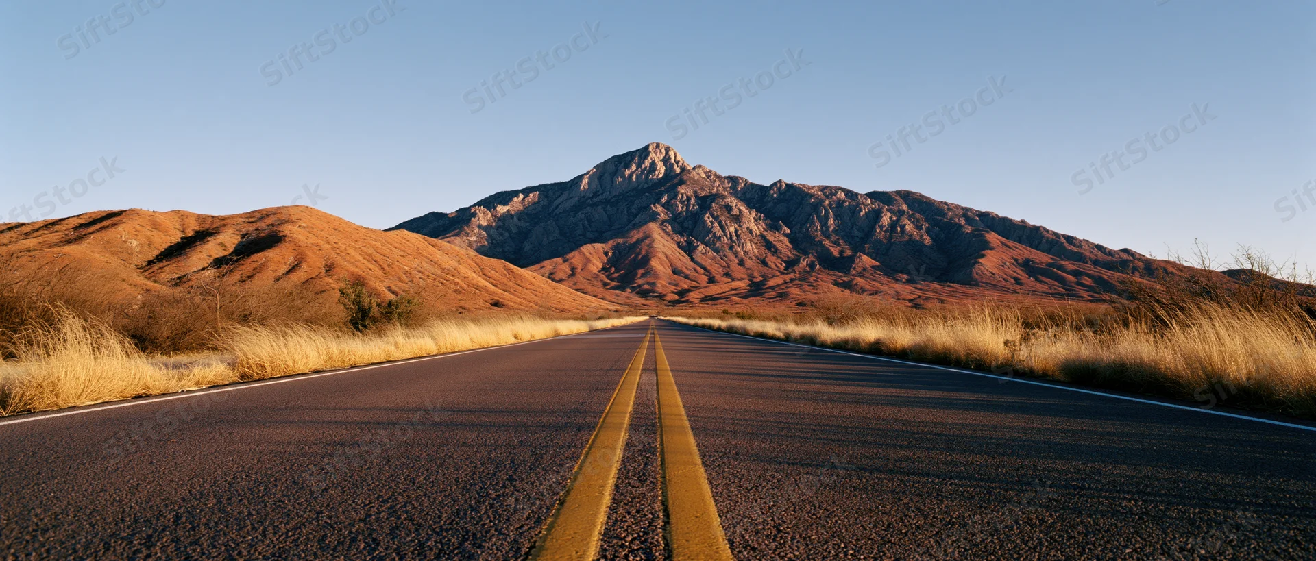 Straight asphalt road with double yellow lines leading toward rocky desert mountains under clear blue sky