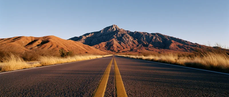 Straight asphalt road with double yellow lines leading toward rocky desert mountains under clear blue sky
