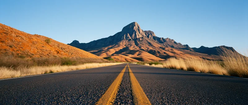 Straight asphalt road with double yellow lines leading toward a rocky mountain in a desert landscape at golden hour