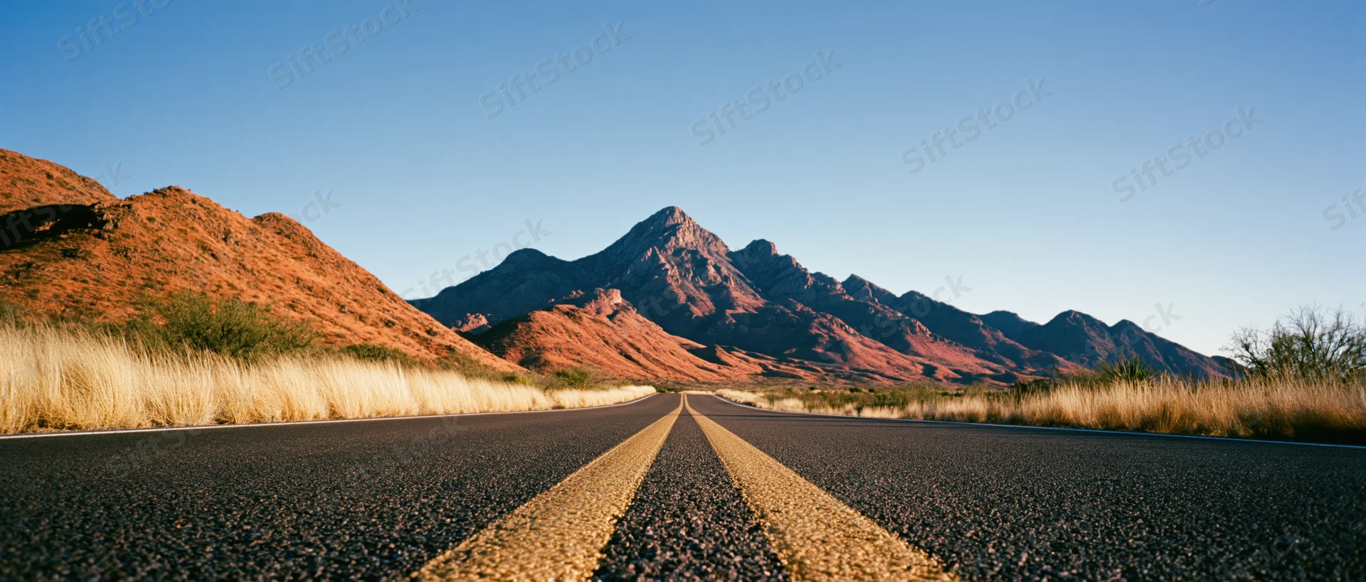 Straight asphalt road leading toward red rock mountains in desert landscape at golden hour