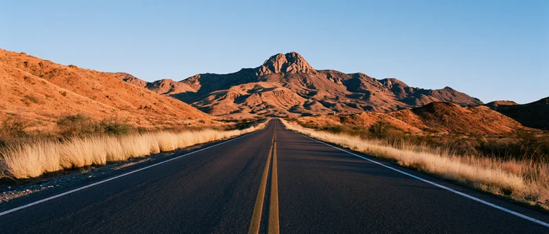 Straight asphalt road leading through arid desert landscape toward rocky mountains at sunset