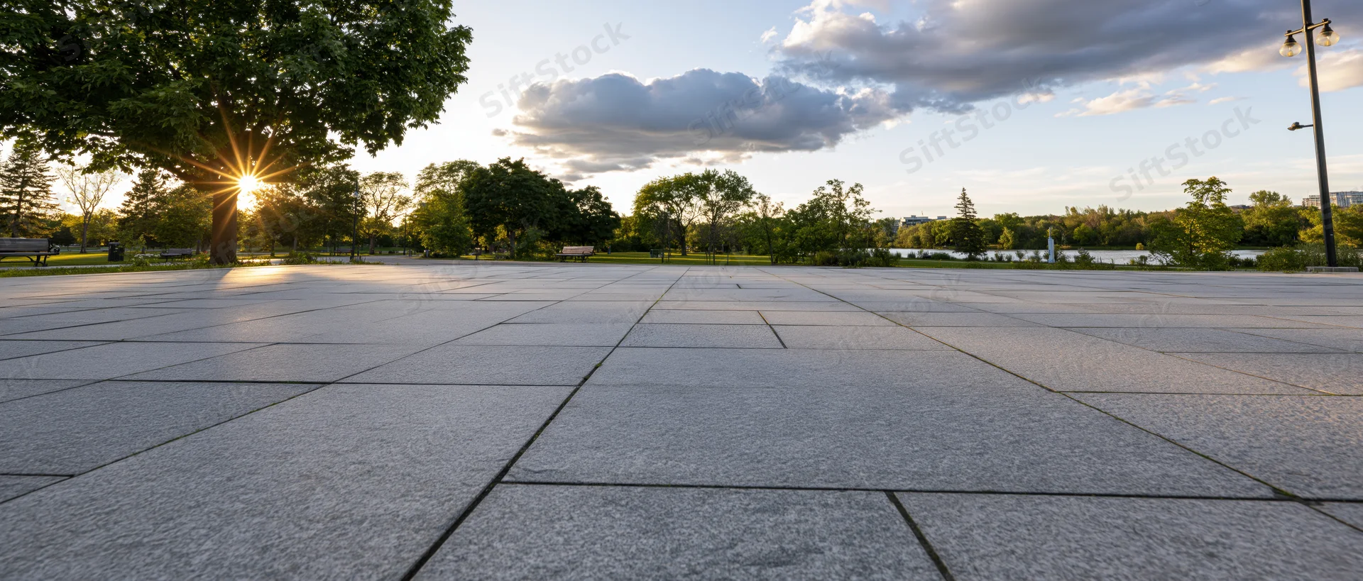 Paved plaza in a park at sunset with trees and river in the background