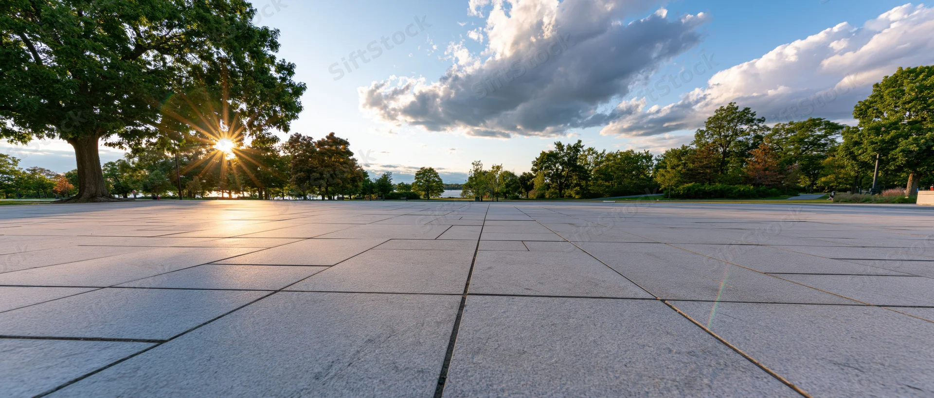 Sunlit stone plaza surrounded by trees under a partly cloudy sky in a park setting
