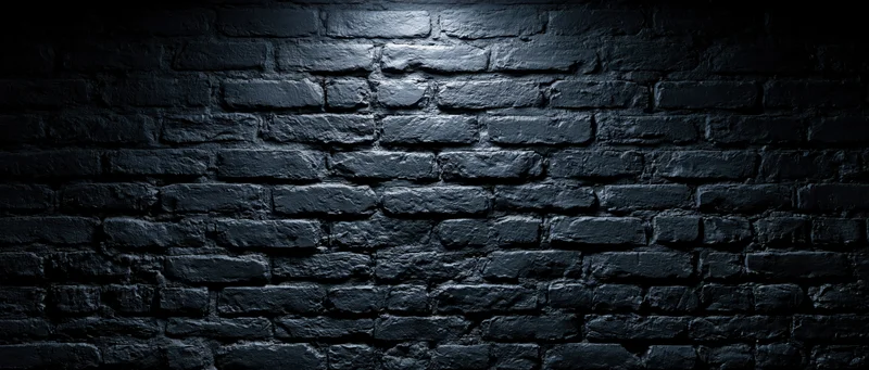 Black brick wall illuminated by spotlight with shadows and texture, creating a dark grunge background with copyspace