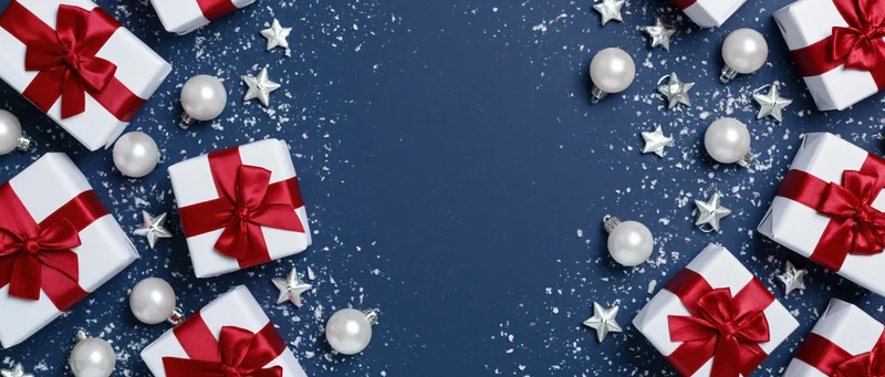 White gift boxes with red ribbons and silver ornaments arranged on blue surface with snow dust and copyspace in center