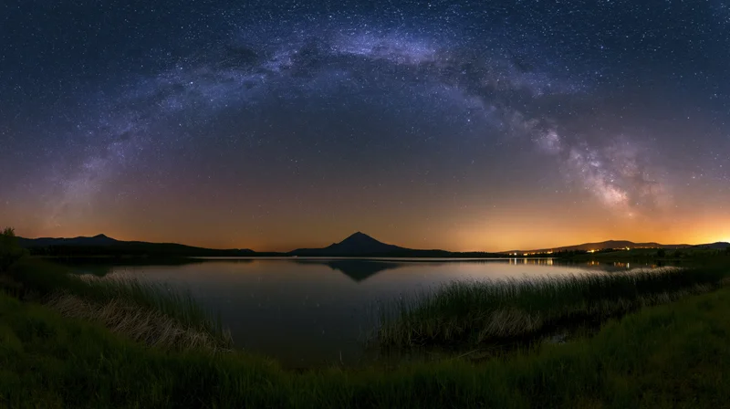 Milky way arching over a calm lake reflecting a distant mountain at twilight