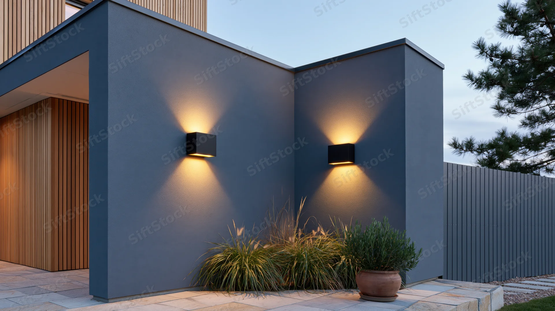 Modern house exterior with wall-mounted sconces illuminating gray facade and garden plants at dusk