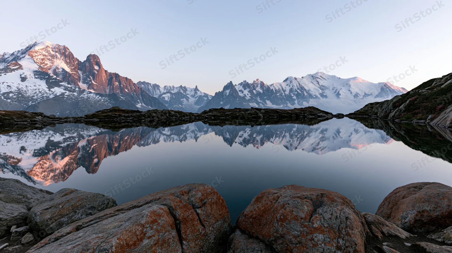 Snow-capped alpine peaks reflected in still lake water at dawn, surrounded by rocky terrain and distant ridges