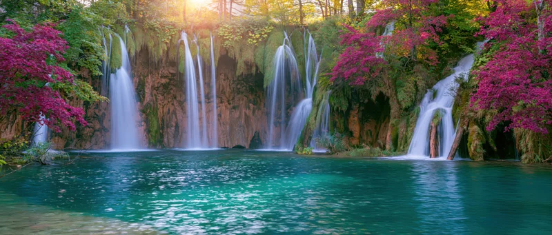 Multiple waterfalls cascading into a turquoise pool surrounded by blooming pink trees and green forest in spring
