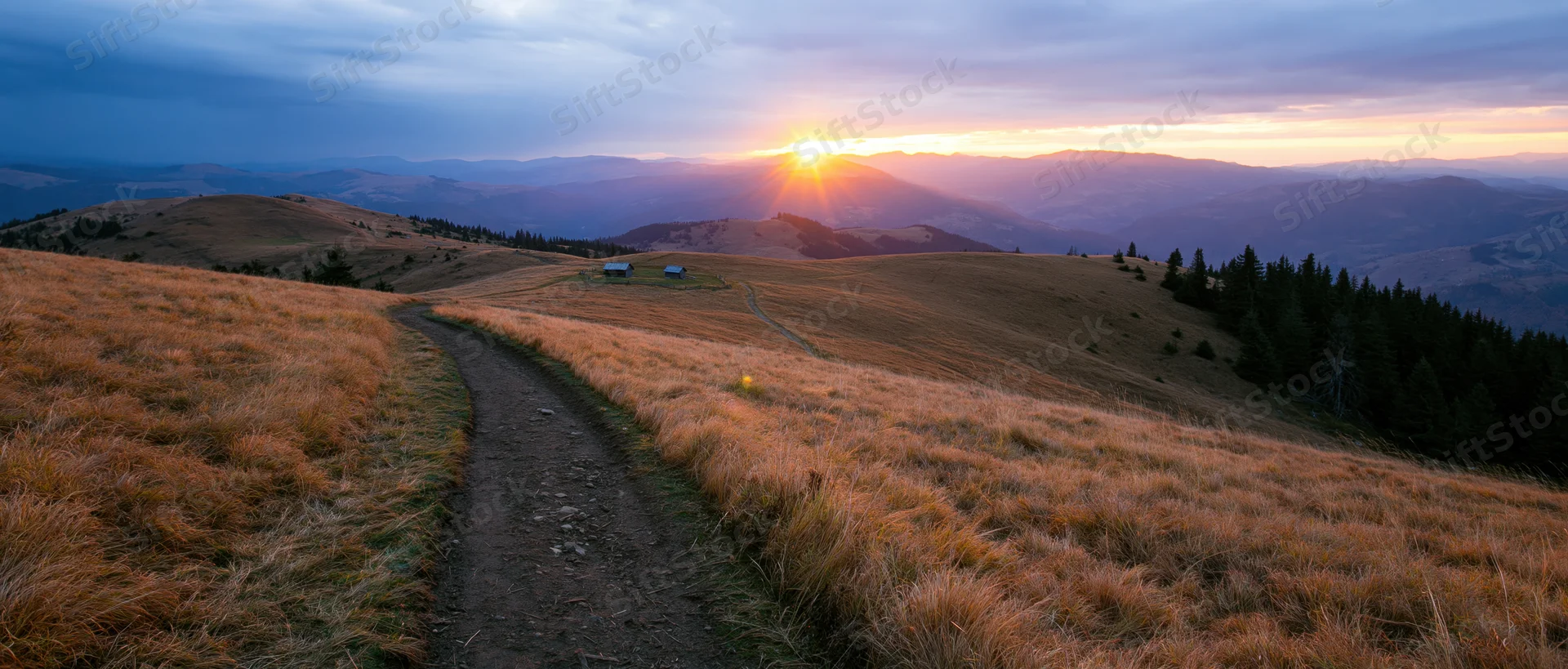 Dirt path winding through golden grassy hills at sunrise with distant mountains and cabins in the valley
