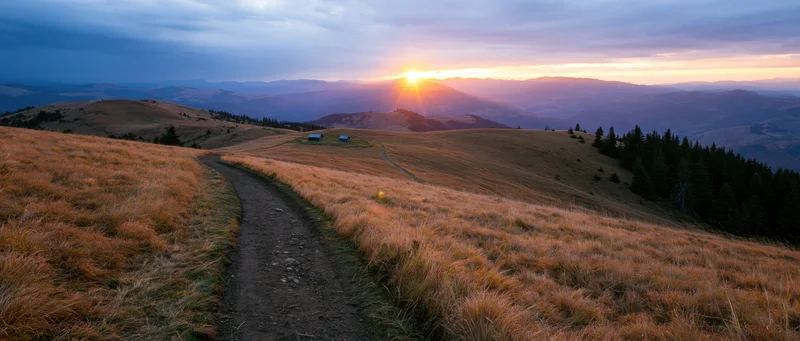 Dirt path winding through golden grassy hills at sunrise with distant mountains and cabins in the valley