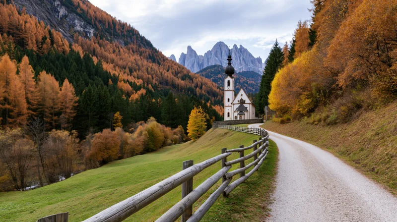 Small white church on a hillside surrounded by autumn trees and mountains, viewed from a gravel road with wooden fence along the side