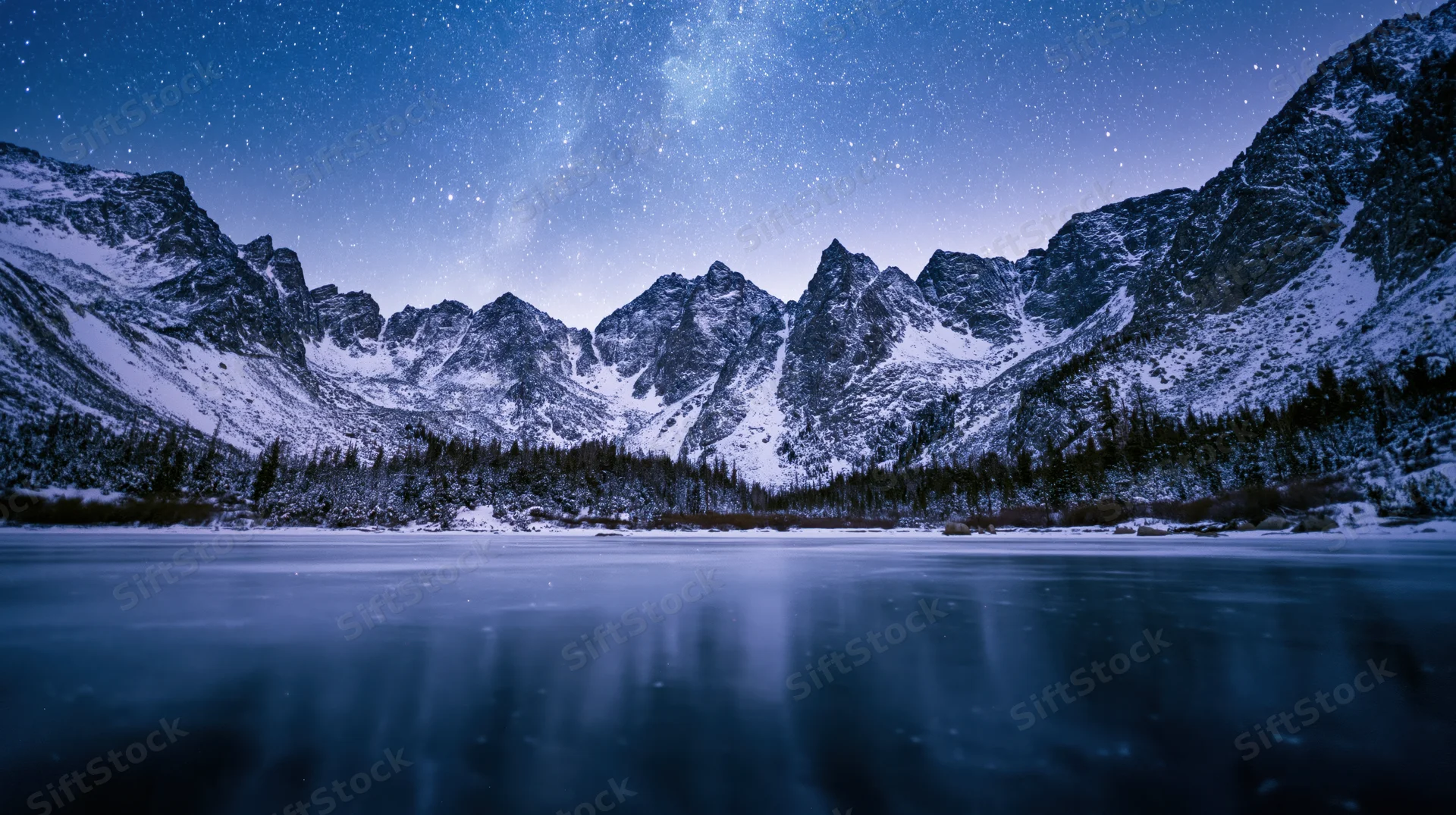 Snow-covered mountain peaks under a starry night sky reflected in a frozen lake