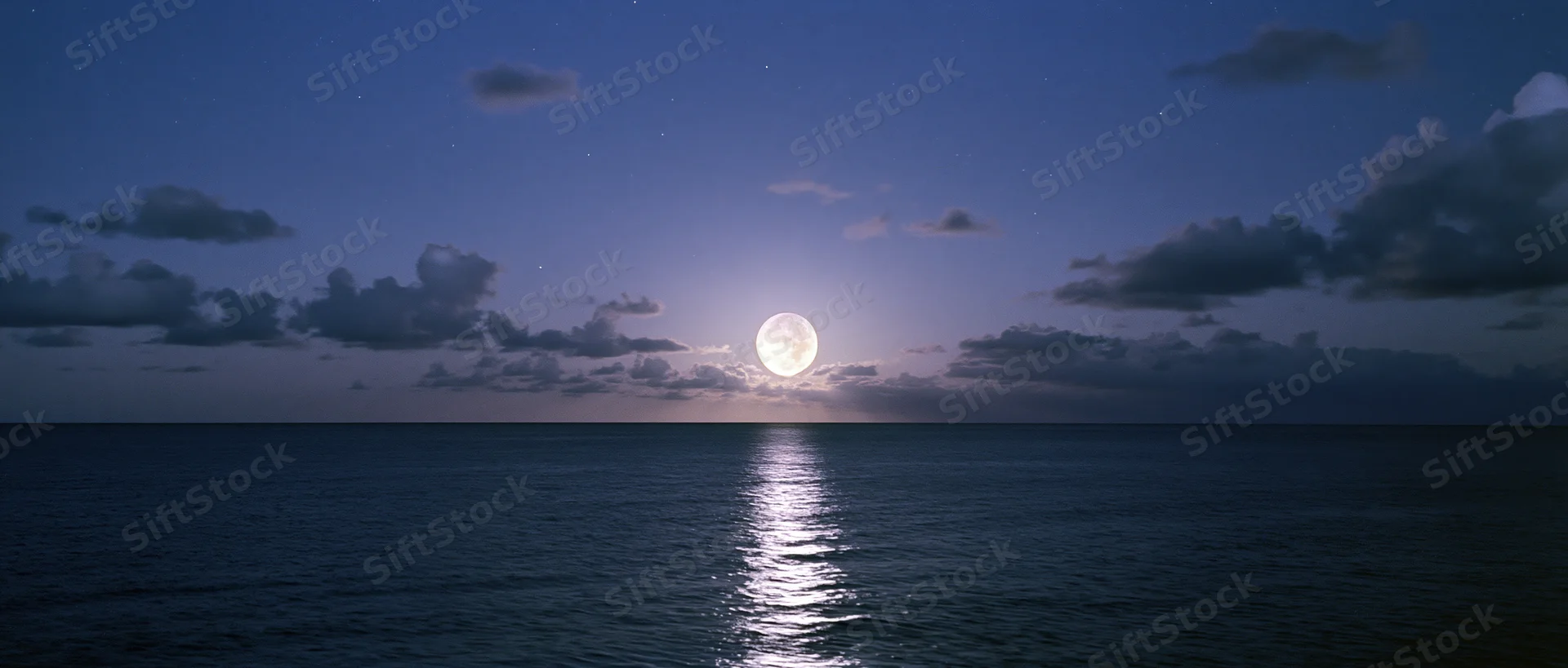 Full moon rising over calm ocean at night with reflection on water and scattered clouds in sky