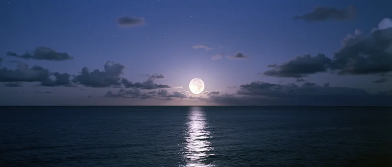 Full moon rising over calm ocean at night with reflection on water and scattered clouds in sky