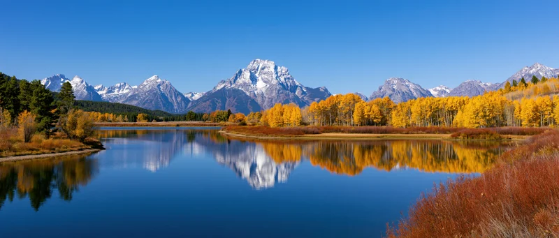 Snow-capped mountains reflected in a calm lake surrounded by autumn aspen trees and evergreens in a mountain valley