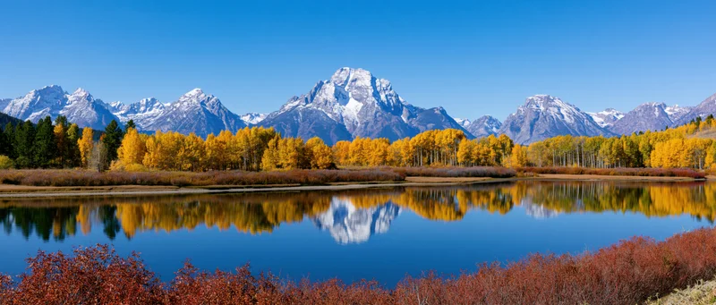 Snow-capped mountain range reflected in calm lake surrounded by autumn aspen trees and evergreens in alpine valley