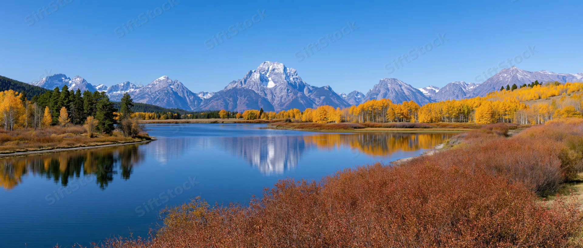 Snow-capped mountain range reflected in calm lake surrounded by autumn aspen trees and evergreens in valley