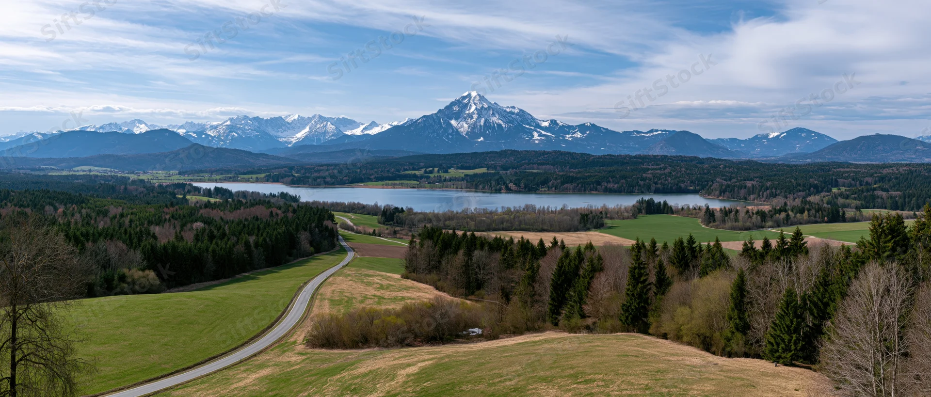 Winding road through green fields and forests leading to a lake with snow-capped mountains in the distance