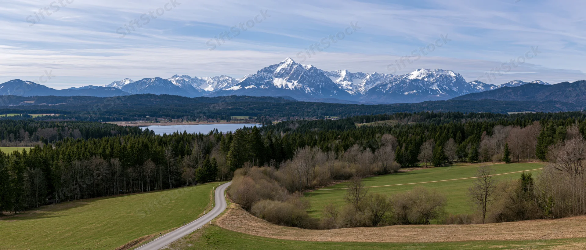 Winding road through green fields and forest leading toward snow-capped mountains in the distance